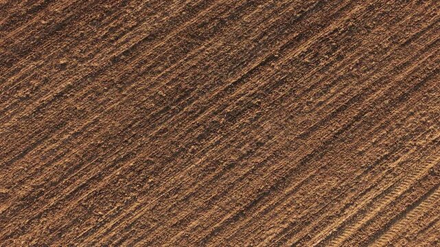 Close aerial view of tilled field with fine parallel ridges and compacted machinery marks across soil. Evenly worked farmland surface ready for seeding with clear texture and natural earth tones