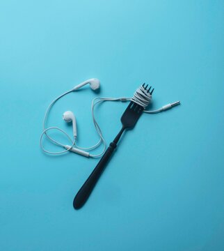 Top view of a creative conceptual layout featuring a matte black fork with a white earphone cable wrapped around it like spaghetti. Minimalist surreal art composition for digital consumption