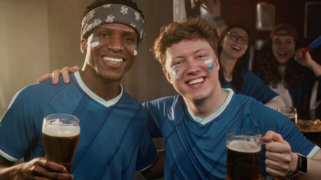 Tilt up slowmo portrait of smiling young diverse male friends with sports fan painted faces wearing team jersey toasting beer glasses in bar with girlfriends cheering in background