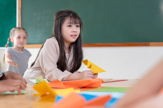 Asian girl playing with colorful paper plane in classroom, Creative elementary students making paper crafts at school desk, Happy children enjoying origami activity in primary classroom
