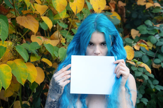 Blue haired woman holding blank white card in front of autumn foliage creating a mysterious portrait with vivid colors strong gaze and copy space for creative concepts