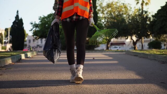 Female municipal worker in reflective safety vest walking through city park carrying rake and garbage bag, preparing for seasonal outdoor maintenance and public space cleaning.