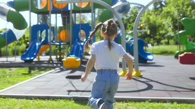 Back view of a happy child running toward swings on park playground.