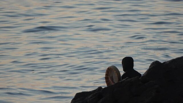 Shaman drumming ocean silhouette during mystic sunset ritual on rocks by the flowing water for spiritual meditation.