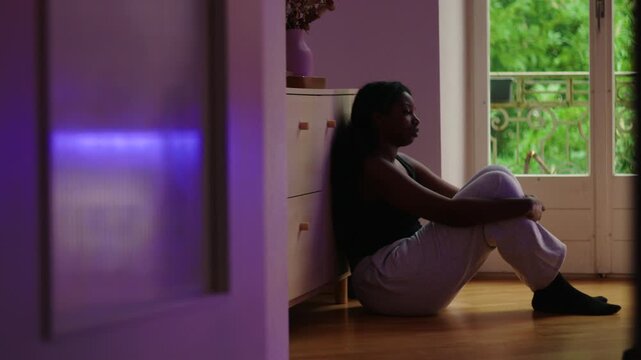 African American woman wide shot sitting on floor by window leaning against cabinet in still posture showing introspection worry and aimless reflection in quiet home space