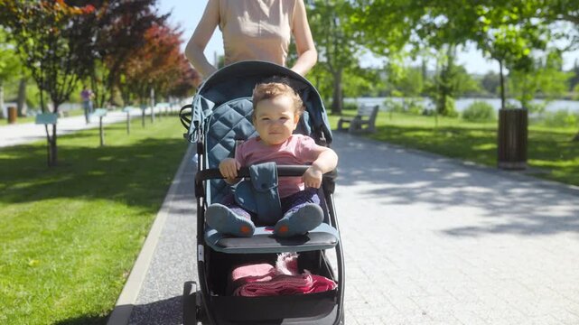 Mother pushing her cute toddler boy in a stroller down a park pathway.