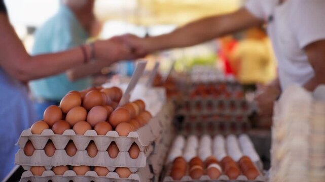 Customer buying fresh brown eggs from a farmer at an outdoor local market stall, with a close-up on cardboard egg cartons and a blurred background of people shopping