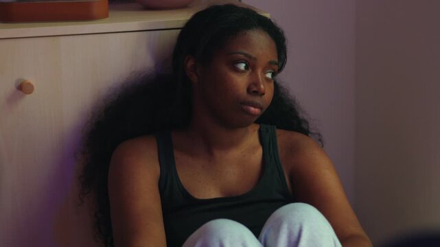 African American woman sitting on floor leaning against cabinet looking aside with distant expression suggesting dissatisfaction contemplation and emotional disengagement in home interior
