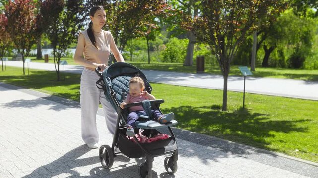 Mother pushing her cute baby boy in a stroller on a sunny park walkway.