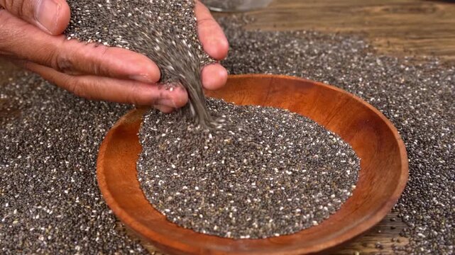 Hands pouring raw chia seeds into a wooden bowl on a textured surface
