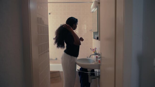 African American woman standing in bathroom looking at herself in mirror with calm expression during quiet self reflection moment in home interior