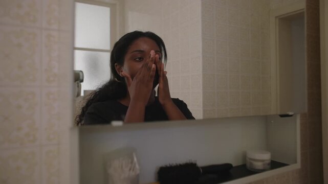 African American woman spreading facial cream across skin then smiling at her reflection in bathroom mirror creating calm and satisfying self care moment in home interior