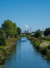 Fototapeta premium Calm blue waterway flows past lush green banks toward distant industrial buildings