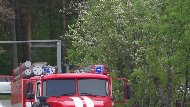 Red fire engine with flashing blue warning light driving near green forest. Emergency vehicle responding to incident. Equipment for fire rescue activity in nature.