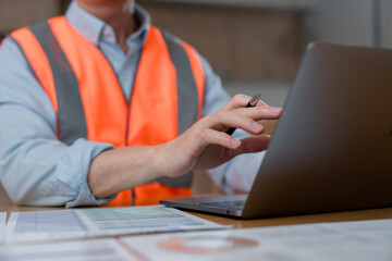Close-up of an industrial worker wearing high visibility safety vest using laptop computer and reviewing documents in office or construction workplace environment. © Teelapon