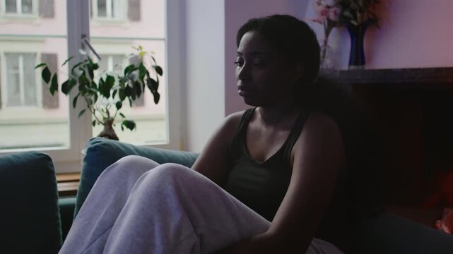 African American woman sitting on couch near window with withdrawn posture, reflecting in silence with subdued sadness, emotional fatigue, and introspective mood in quiet home interior