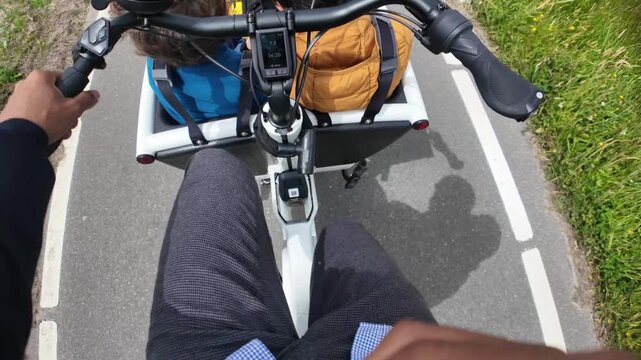 Father riding an electric cargo bike with his two sons on a bike path, showcasing sustainable family commuting, pov shot with hands on the handlebars and legs pedaling