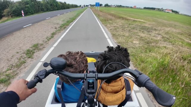 First-person view of a father riding an electric cargo bike with his two young sons sitting in the front on a dedicated cycle path through the countryside