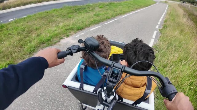 Father riding an electric cargo bike with his two children in the front, enjoying a family outing on a paved cycle path through the countryside with a first-person view