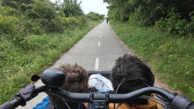 Point of view shot of a cyclist riding an electric cargo bike on a paved path in the countryside with two children in the front basket, enjoying a family outing together