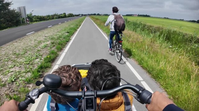 First-person view of a family riding an electric cargo bike and a regular bicycle on a dedicated bike path through the green countryside on a cloudy summer day