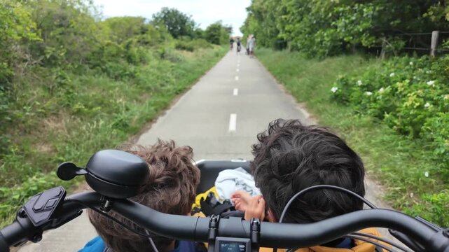 First-person view of a cargo bike ride with two children in the front basket, cycling along a narrow asphalt path surrounded by lush green summer vegetation and wildflowers