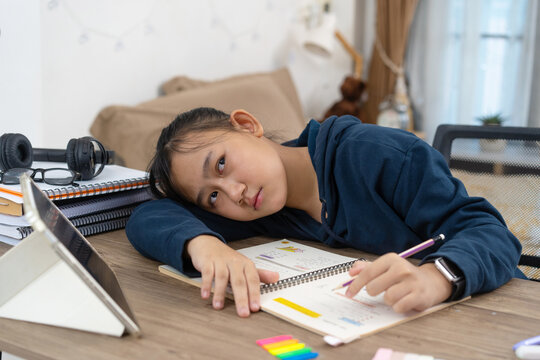 Tired young girl rests her head on the desk with notebooks, headphones, tablet while studying at home