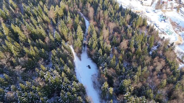 Solar powered vehicle parked in a snowy forest as drone tilts up from steep downward view, revealing conifer woodland landscape in Parc naturel r&eacute;gional du Haut Jura, France.
