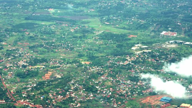 A peaceful window-seat perspective capturing vibrant green fields and rural villages in Uganda, with light clouds passing below the aircraft near Entebbe.