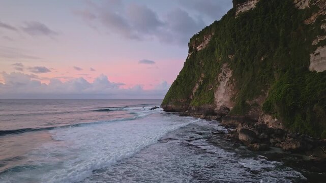 Lush green cliffside meeting the Indian Ocean at Green Bowl Beach, Bali, showcasing waves breaking on the shore under a beautiful pink and purple twilight sky. Static shot