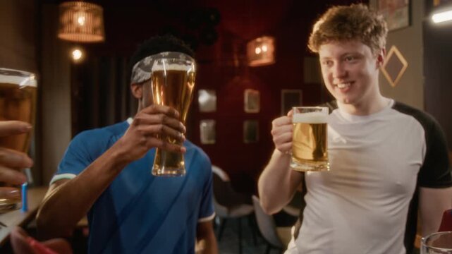 Slowmo shot of joyful young diverse friends in team jersey sitting at bar table with snacks and raising beer glasses while celebrating sports win