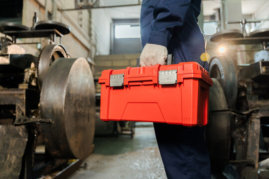 Close up of an industrial mechanic hand holding a red plastic toolbox container while standing near heavy machinery equipment for a repair service inside a traditional factory workshop