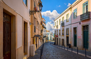 Obraz premium Lisbon, Portugal. Antique Alfama district with traditional portugese houses on background of blue sky with clouds. Paving stones narrow street between buildings. Decorative street lamp on the wall.