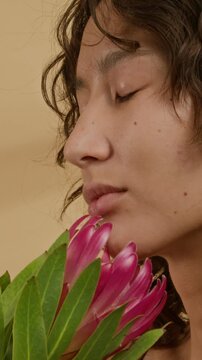 Vertical close-up shot of gen Z girl with curly hair enjoying smell of pink protea flower with closed eyes against studio background