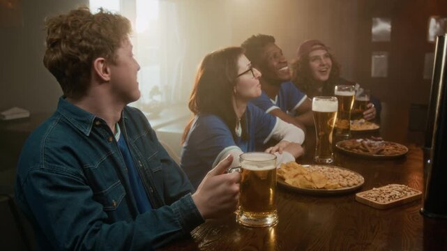 Side view shot of cheerful young multiethnic group of friends watching live sports while sitting at bar counter with assorted snacks and beer glasses, cheering and having fun together