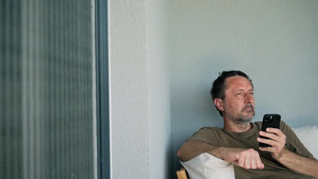 Tired middle-aged man sits on balcony sofa, yawning and covering his mouth while checking his smartphone, conveying fatigue, boredom, doomscrolling and everyday digital life. 4K.