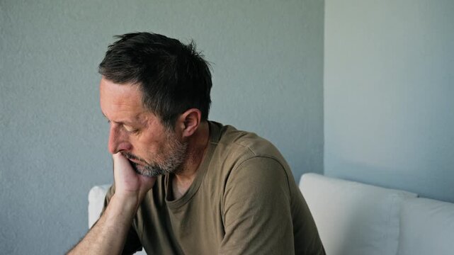 Middle-aged man sits on a sofa with his hand on his chin, appearing pensive and worried. 4K