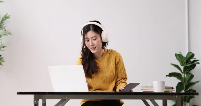 Young asian woman wearing headphones while diligently writing in a notebook, concentrating on her studies at home, engaged in self-learning and remote work with technology