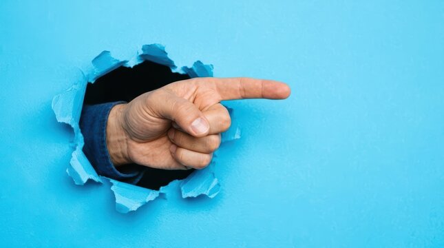 Hand pointing through torn paper hole into blue background, finger extended from dark interior opening in studio backdrop
