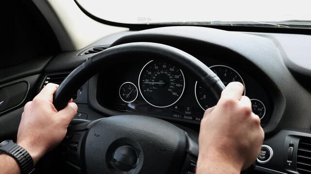Male arms holds steering wheel while driving a motor car at city. Young man riding on electric automobile at urban road. Driver moving on his electrical auto on route. Slow motion