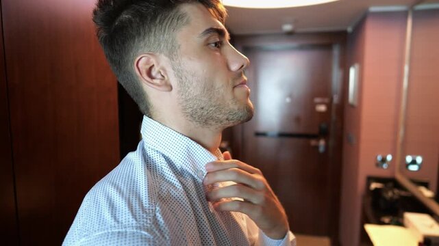 Handsome businessman adjusting shirt collar in front of mirror in modern hotel room or apartment interior. Young manager preparing for business meeting or formal event showing confidence. Close up