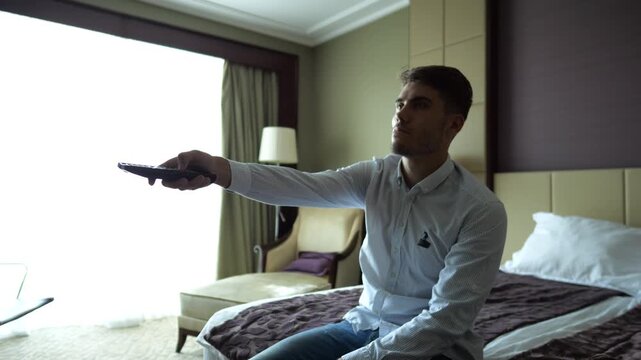 Young businessman switch channels on TV remote control sitting on bed in modern hotel room after travel. Relaxed man enjoying leisure during business trip in comfortable hospitality interior. Close up