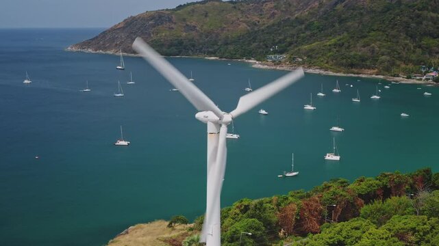 Rotating wind turbine blades generating renewable energy on hill overlooking a peaceful bay filled with anchored sailboats and yachts nestled against coastline of Phuket island. Aerial view drone shot