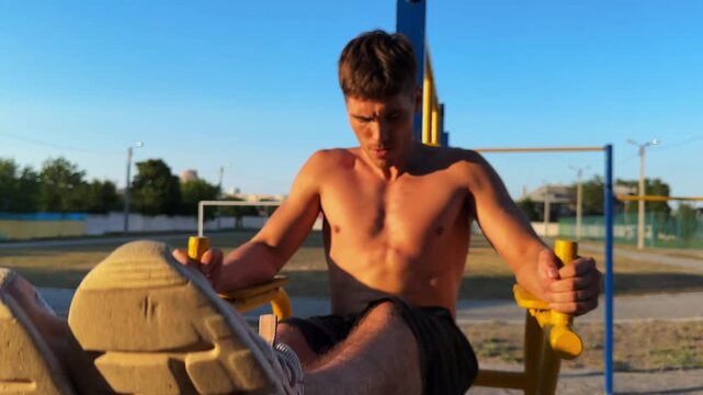 Group of young shirtless sportsman trains abdominal muscles raising legs on parallel bars at sports ground. Strong man exercising at nature. Sporty athlete working out at nature. Sportive lifestyle