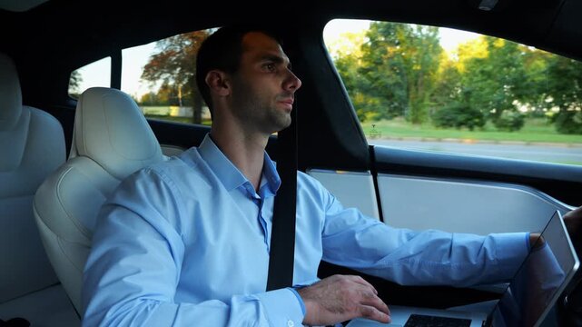 Male businessman typing text on laptop during riding on electrical vehicle with autopilot. Handsome entrepreneur working on notebook while riding an autonomous self driving electric car at urban road