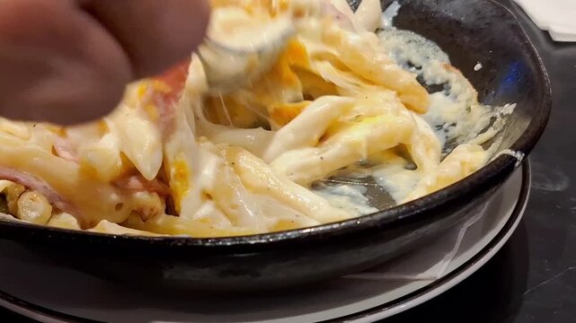 Close-up of a spoon lifting delicious baked penne pasta covered in creamy white sauce and melted, stretched mozzarella cheese from a hot black pan or iron skillet.
