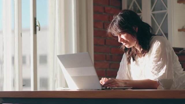 Portrait of a young woman working on a laptop at a kitchen table. Remote work, home office, focused professional, modern lifestyle, productivity in a cozy home interior.