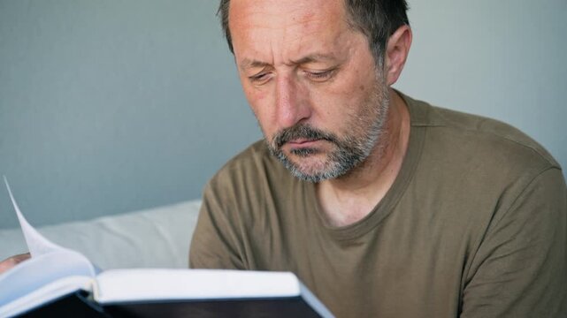 Contemplative middle-aged man reading a book and thinking deeply at home, 4K
