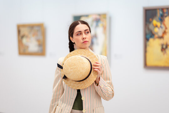 Portrait of a young Caucasian pensive woman elegant jacket and trousers holds straw hat and posing in the art gallery. Painting in background. Concept of cultural education and Museum Day