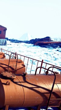 Containers sit on a platform by the ocean. Waves crash against the rocks. The sun shines brightly, and a distant landform is visible. The scene shows a busy coastal area.